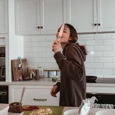 a woman brushing her teeth while standing in a kitchen with white cabinets and counter tops