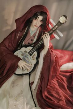 a doll is holding a guitar and wearing a red cloak while sitting on a bed