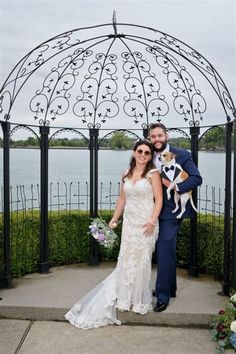 a bride and groom pose with their dog in front of an iron gazebo by the water