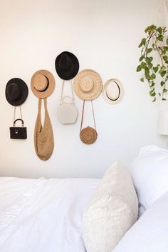 hats and purses hanging on the wall next to a bed with white linen pillows