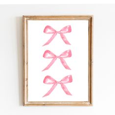 three pink bows hanging on the wall in a wooden frame against a white wall with wood trim