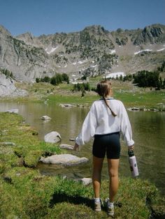 a woman in shorts and a white shirt is walking towards a lake with mountains in the background