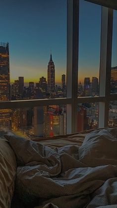 an unmade bed in front of a large window overlooking the city at night time