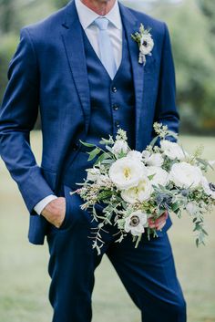 a man in a blue suit holding a bouquet of white flowers and greenery on his lapel