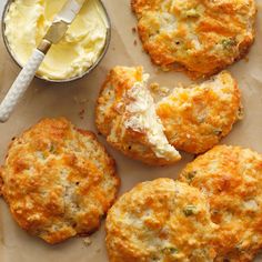 four biscuits with cheese and butter next to a bowl of whipped cream on a piece of parchment paper