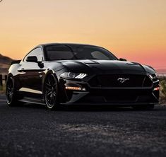 a black mustang parked on the side of a road at sunset with its lights on