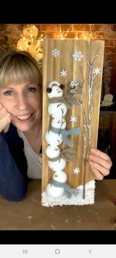 a woman is holding up a snowman and tree decoration on a wooden board with lights in the background