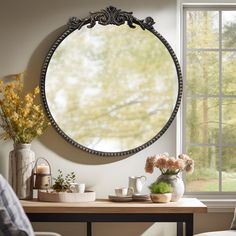 a round mirror hanging on the wall next to a table with flowers in vases