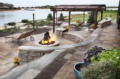 an outdoor fire pit with benches and chairs around it, near the water's edge