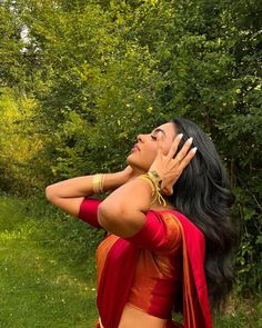 a woman in a red and gold sari with her hands on her face looking up into the sky