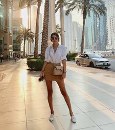 a woman is standing on the sidewalk in front of palm trees and buildings with her purse