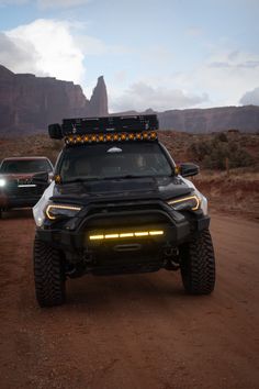 the front end of a truck with lights on driving down a dirt road near mountains