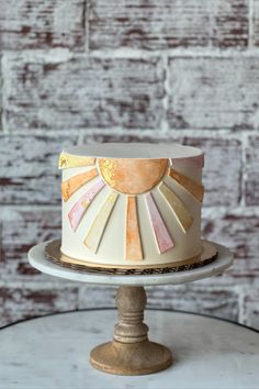 a white cake with gold and pink decorations on a stand next to a brick wall