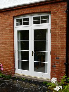 a brick building with two white double doors and flowers in the foreground, next to an open patio door