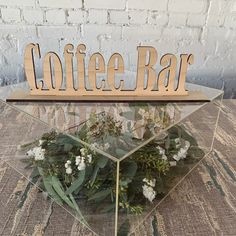 a coffee bar sign sitting on top of a glass shelf filled with flowers and greenery