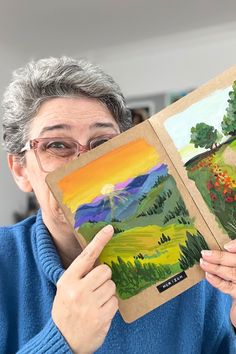 an older woman holding up two paintings in front of her face