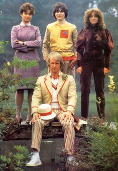 three men and one woman are sitting on a bench in front of some flowers,