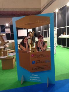 two women sitting in a cardboard box on display at an expo or exhibition with green carpet and blue walls