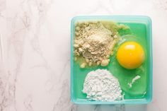 an egg, flour and other ingredients in a green container on a marble countertop