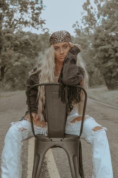 a woman sitting on top of a metal chair in the middle of a road with trees behind her