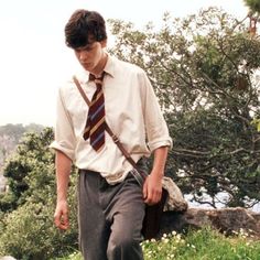 a young man wearing a tie and slacks walking up a hill with trees in the background