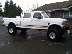 a white pickup truck parked in front of a house with large tires on it's flatbed