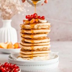 a stack of pancakes with syrup being drizzled over them and topped with cherries