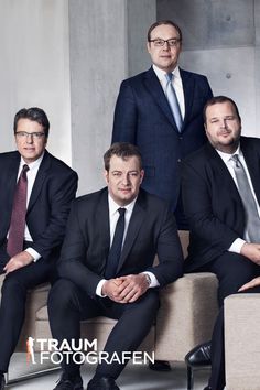 three men in suits and ties are sitting on a couch with their arms crossed, posing for the camera