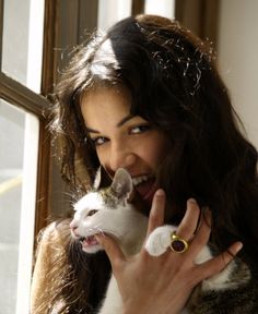 a woman holding a cat in her right hand and smiling at the camera while standing next to a window