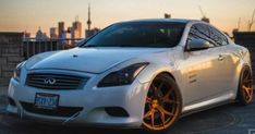 a white car with gold rims parked in front of a cityscape at sunset