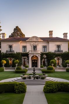 a large white house with lots of trees and bushes in the front yard at sunset