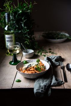 a bowl filled with pasta on top of a wooden table next to a bottle of wine