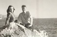 an old photo of two people sitting on a rock by the ocean, looking into the distance