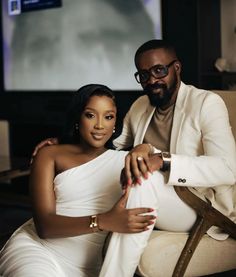 a man and woman sitting on a couch in front of a television screen, posing for the camera