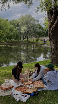 three women sitting on the grass with pizzas in front of them and a lake behind them