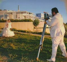 a man in white suit taking pictures with a camera on the grass near a bride and groom