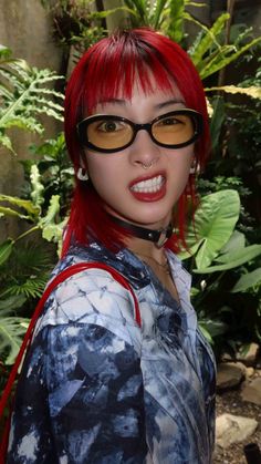 a woman with red hair and glasses posing in front of some plants, wearing sunglasses