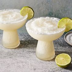 two margarita cocktails with lime slices on the rim and salt in bowls next to them