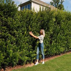 a woman standing in front of a hedge picking something off the ground with her hands