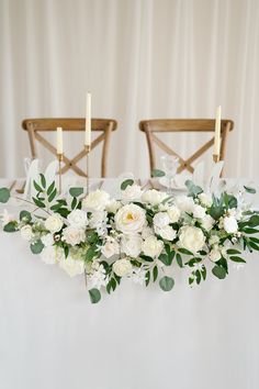 white flowers and greenery are arranged on a table with two chairs in the background