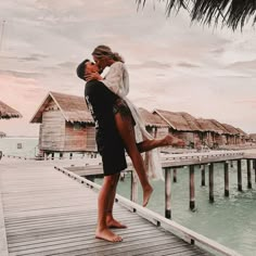 a man and woman kissing on a dock next to water with thatched huts in the background