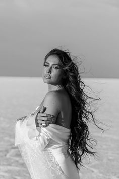 a woman with long hair standing on the beach looking off into the distance while wearing a white dress