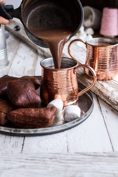 a person pours hot chocolate into two mugs on a plate with marshmallows