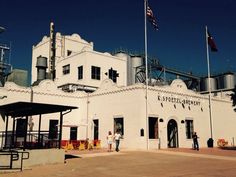 an old white building with flags flying in the wind and people standing outside it on a sunny day