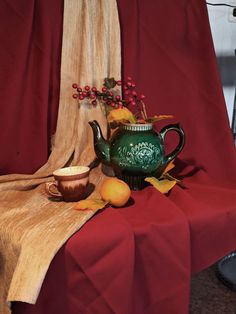 a green teapot and some fruit on a red table cloth next to a curtain