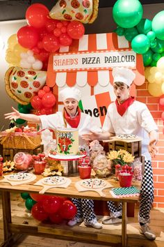 two chefs standing behind a table with pizzas on it and balloons in the background