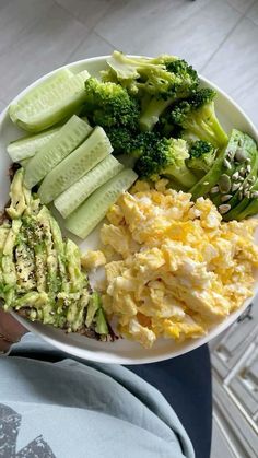 a person holding a plate with eggs, broccoli and cucumbers on it