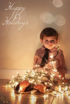 a little boy sitting on the floor next to a christmas tree with lights around him