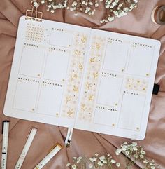 an open planner sitting on top of a bed next to some pens and flowers,