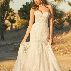 a woman in a wedding dress standing on a dirt road with palm trees behind her